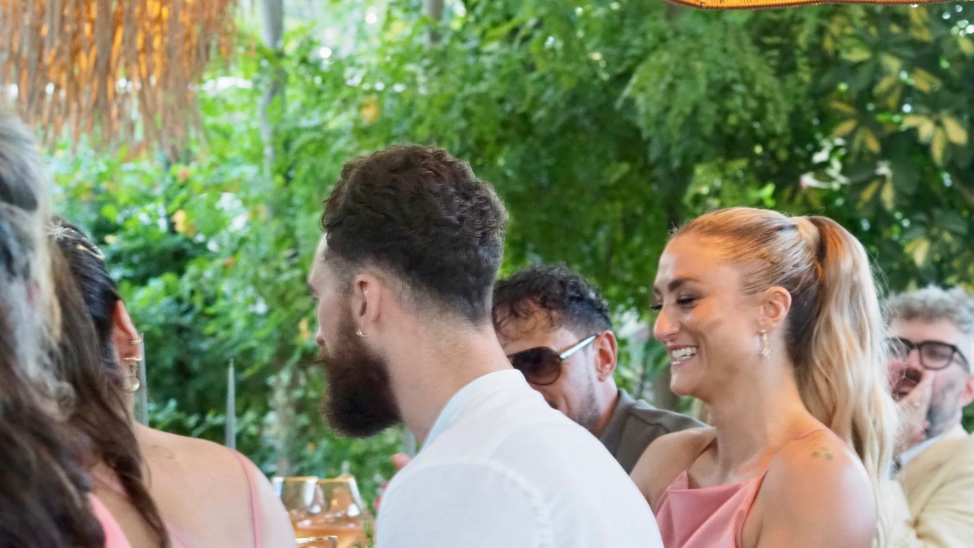 People chatting at an outdoor event, with greenery and a smiling woman in a pink top in the foreground