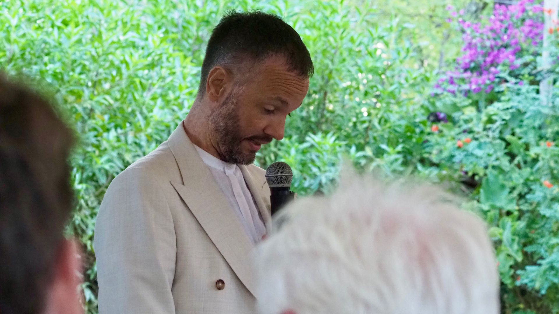 Man in a light suit speaking into a microphone at an outdoor gathering, with greenery and flowers behind him