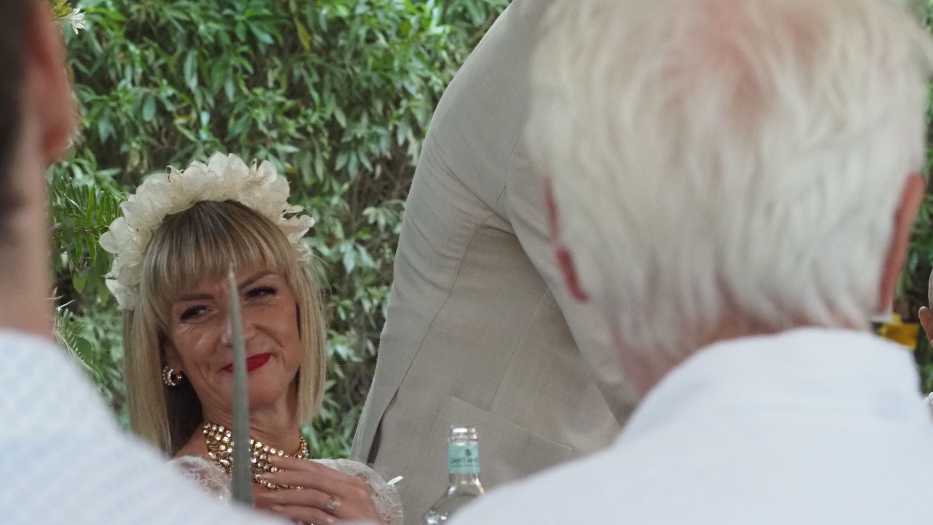 Woman in white floral headpiece seated at a table, smiling in a garden setting during an outdoor gathering