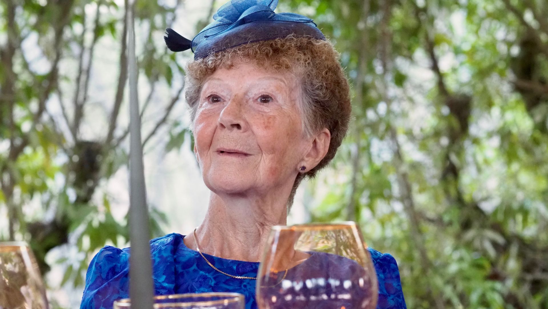 Elderly woman in a blue dress and hat seated outdoors at a table, with a drink in the foreground.