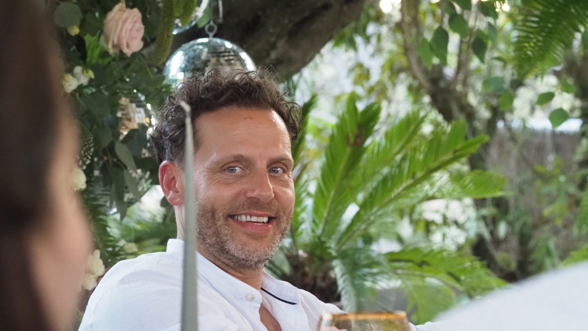 Smiling man in white shirt at an outdoor gathering, framed by greenery and palm leaves