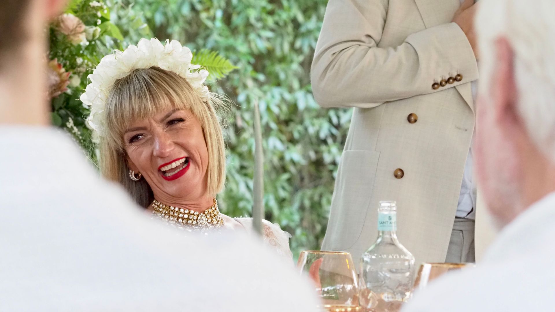 Smiling woman with blonde hair at an outdoor gathering, seated beside a table with drinks and guests nearby