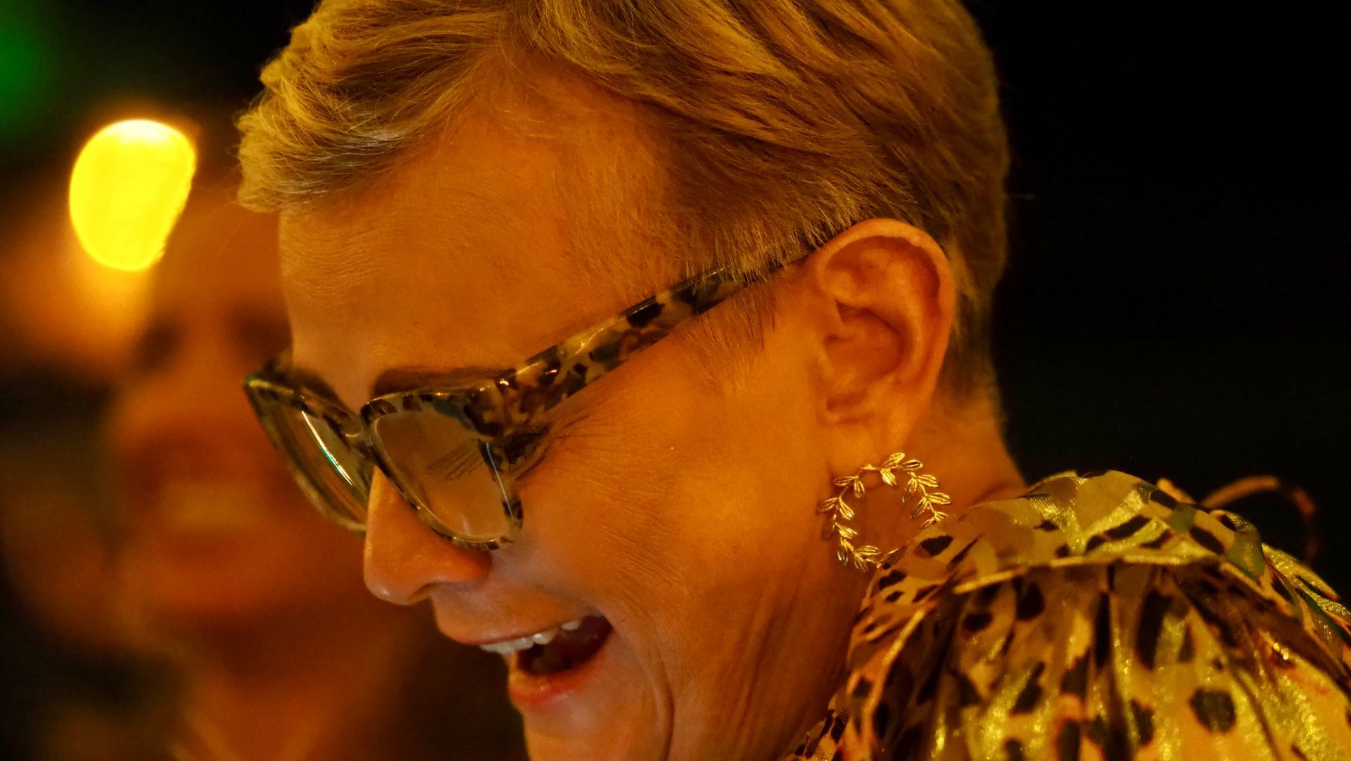 Smiling person with glasses and floral earrings in warm, dim lighting