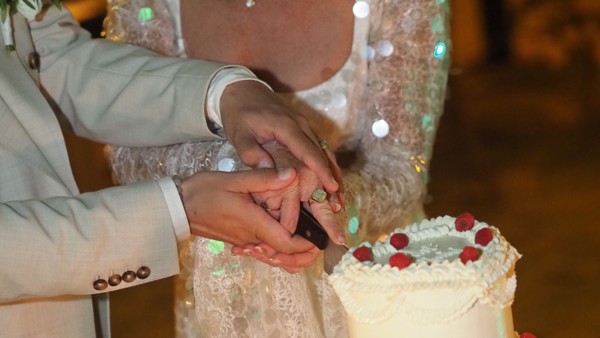Hands cutting a white wedding cake decorated with red and green flowers.