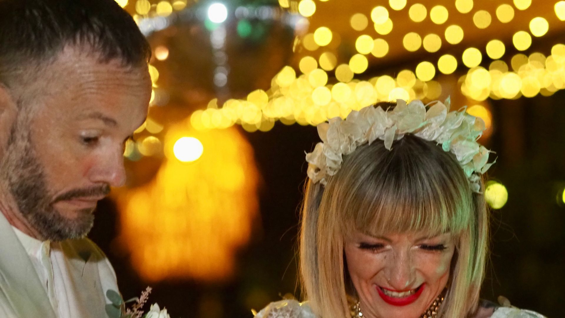 Two people in formal wear smiling under warm wedding lights, with a floral headpiece visible.