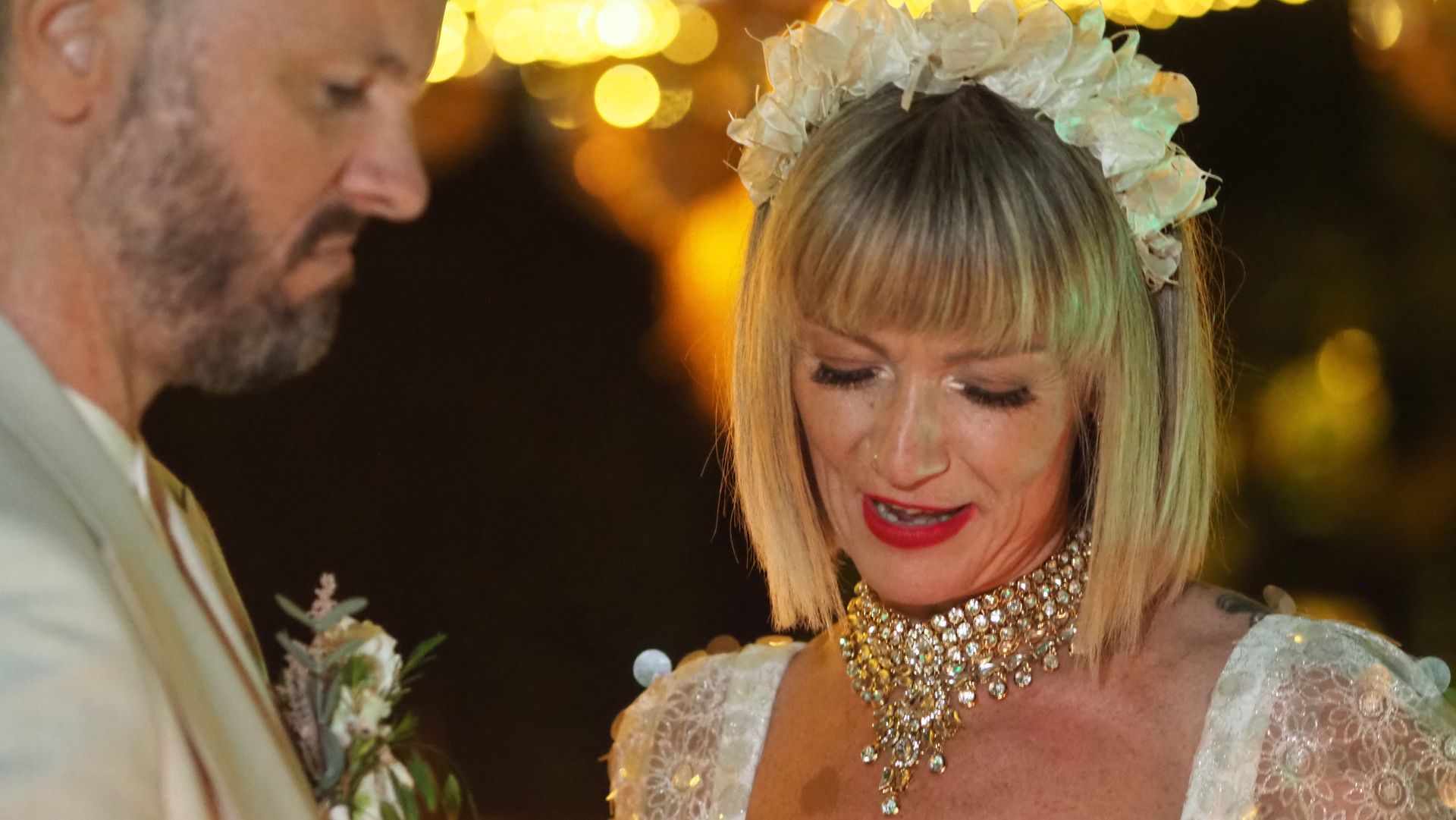 A couple in formal attire at a warmly lit event, with the woman wearing a floral headpiece and jeweled necklace.
