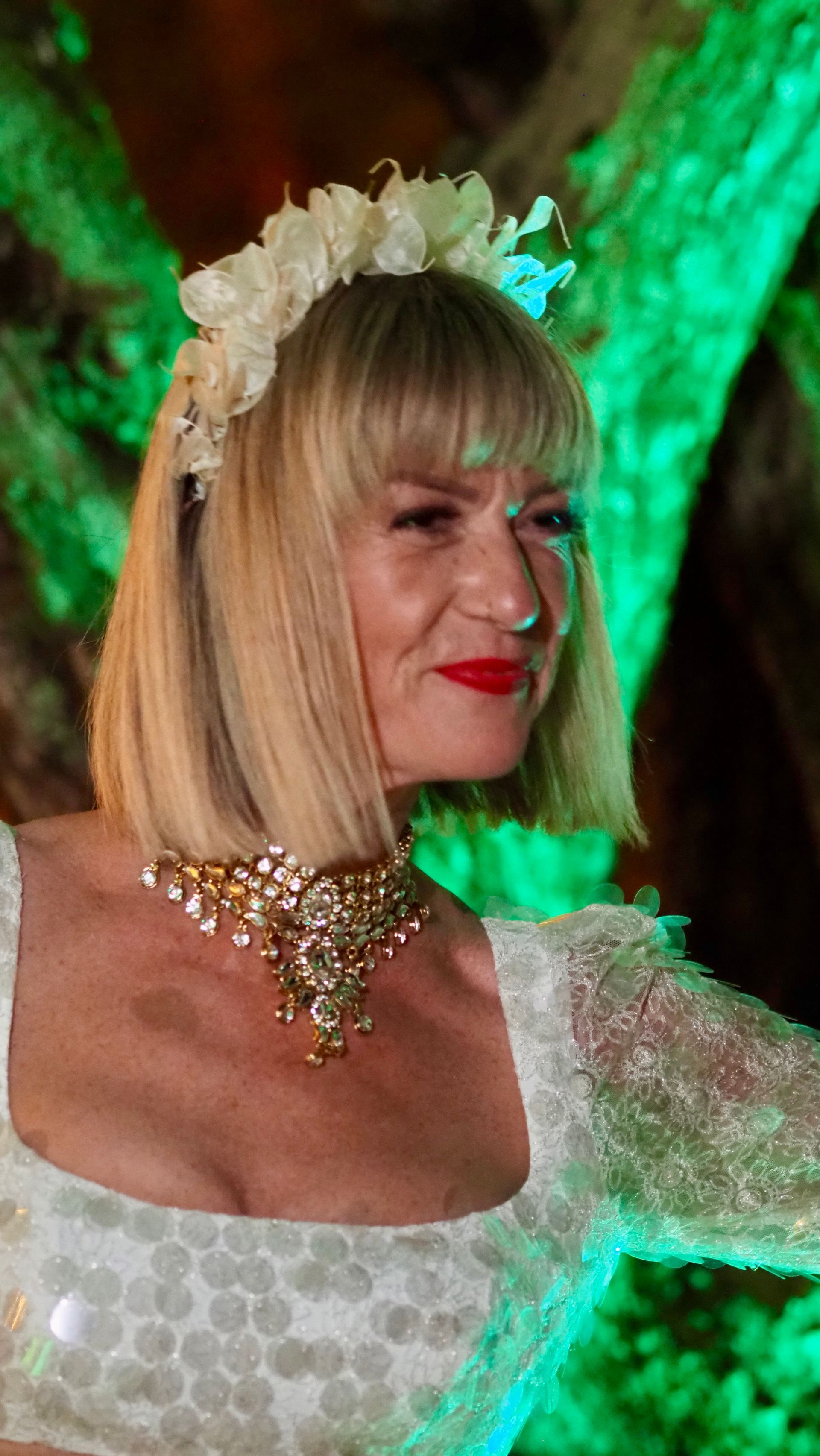 Blonde person in a white dress and floral headpiece, wearing a jeweled necklace, at a green-lit event backdrop