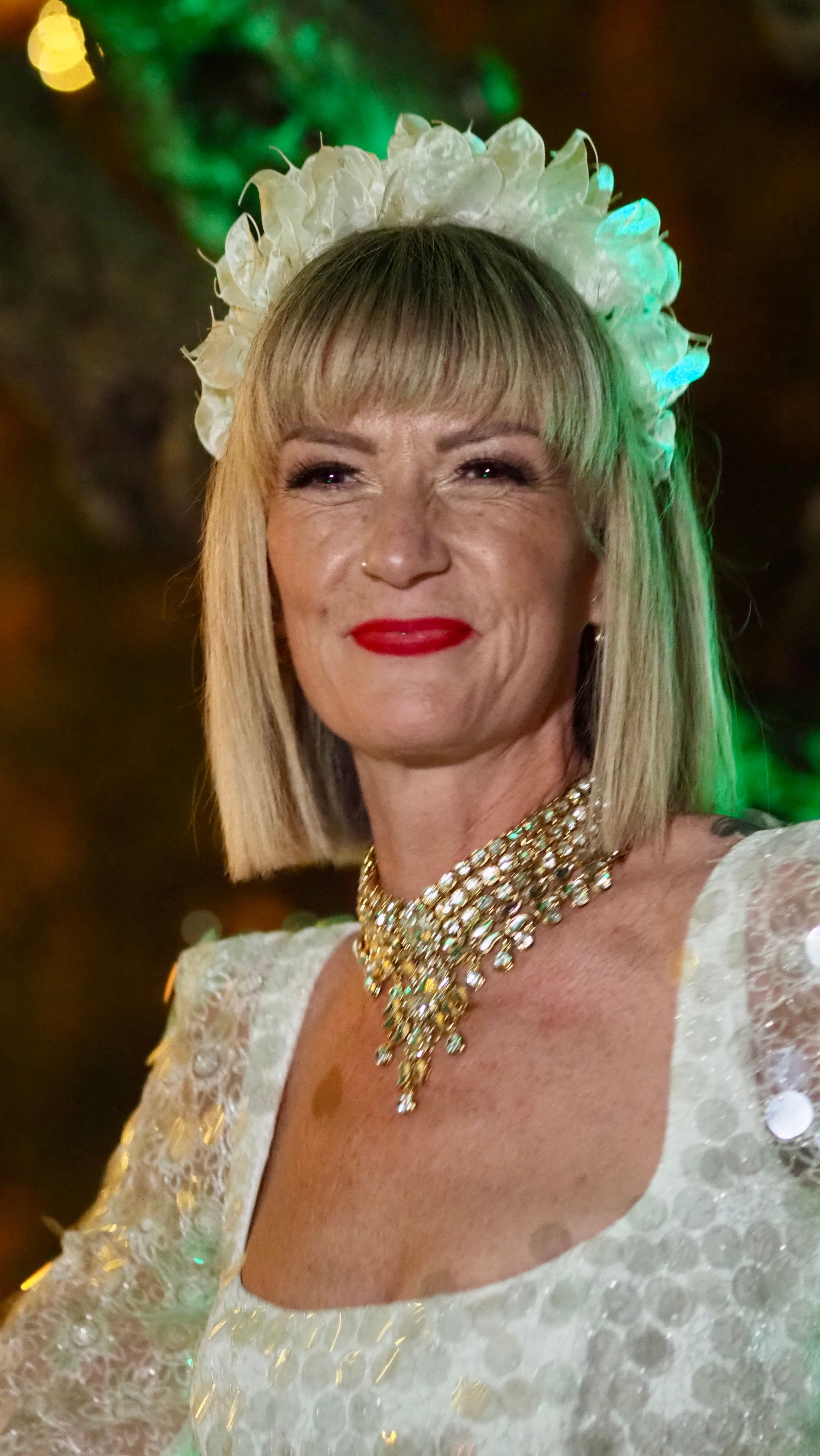 Woman in a white dress and floral headpiece, wearing gold jewelry and red lipstick, posing against a green background