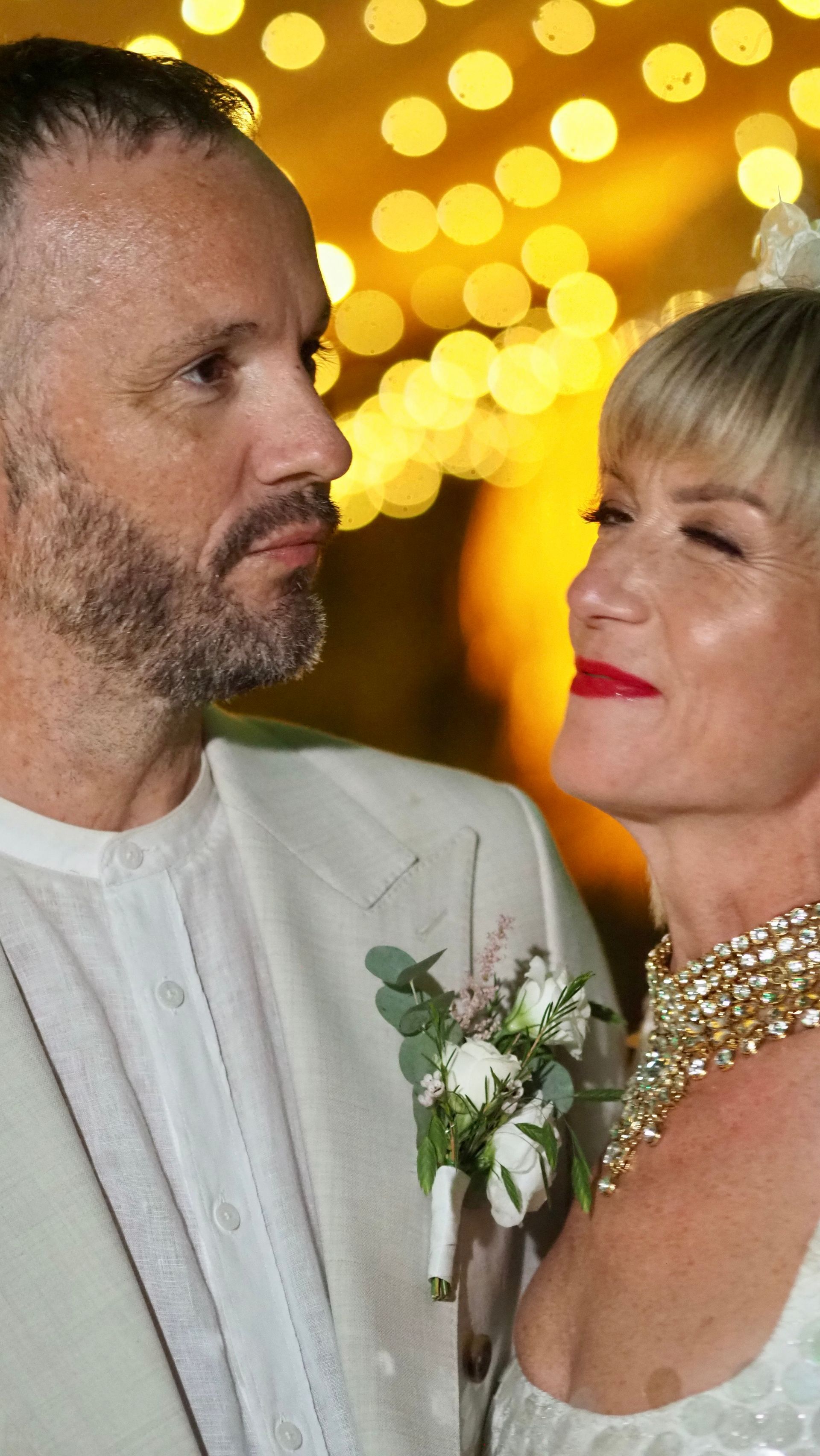Two people in white outfits facing each other at a warm, glowing event backdrop.
