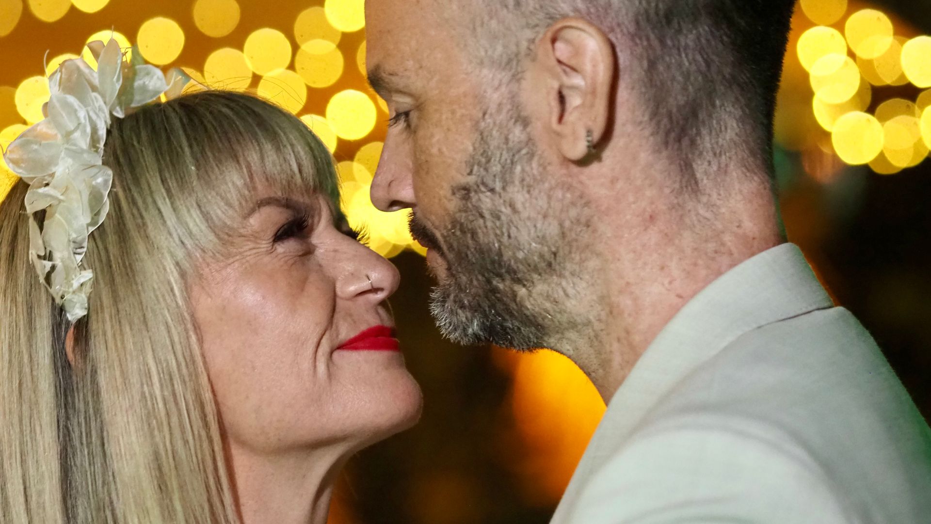 Two people in formal wear face each other closely with warm bokeh lights in the background.