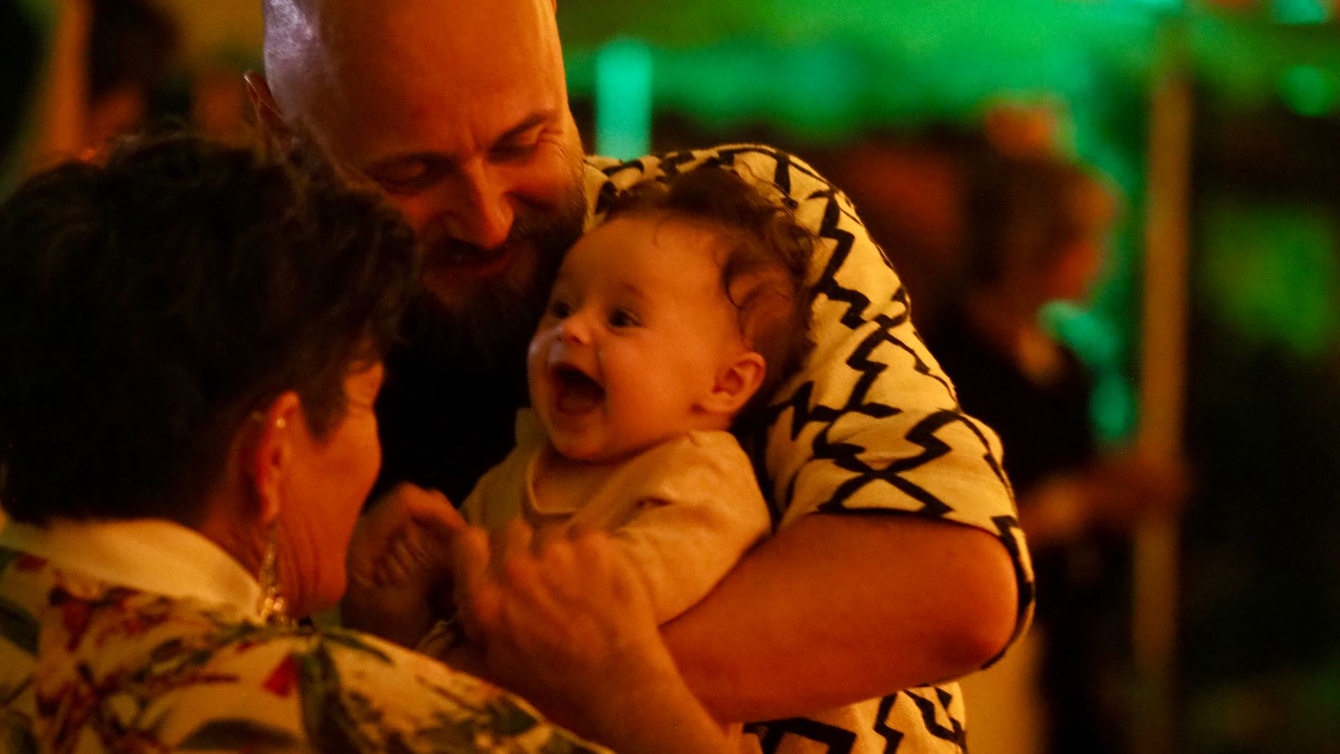 A man smiles and holds a laughing baby in a warmly lit, crowded indoor setting.