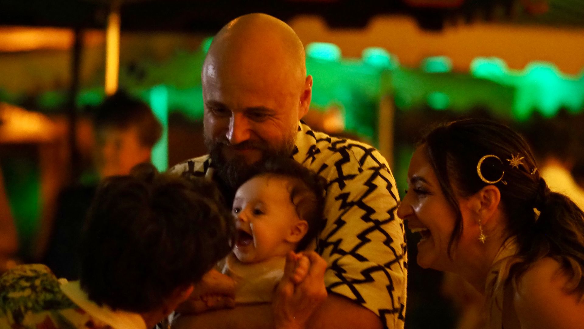People laughing together at a warm-lit event, one holding a smiling baby.