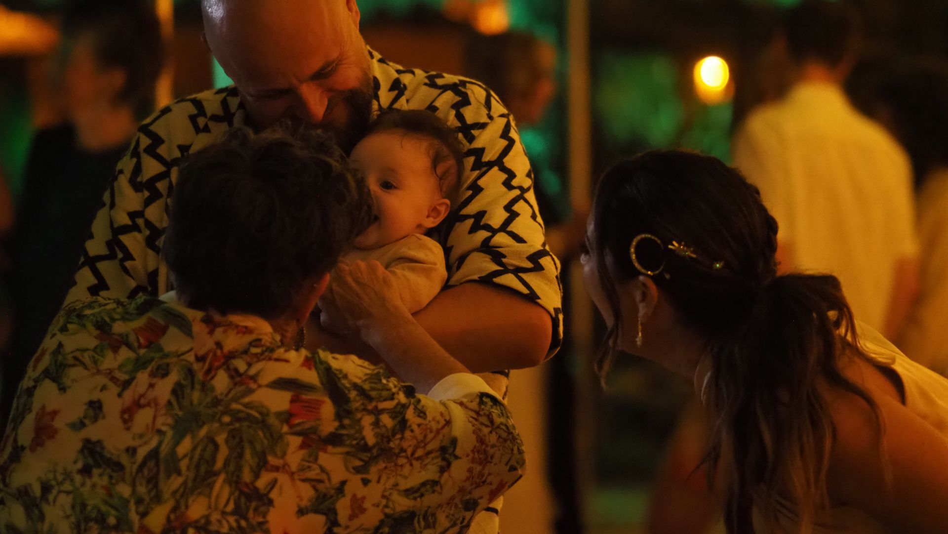 People gathered at a warmly lit outdoor event, with a baby being held in the center.