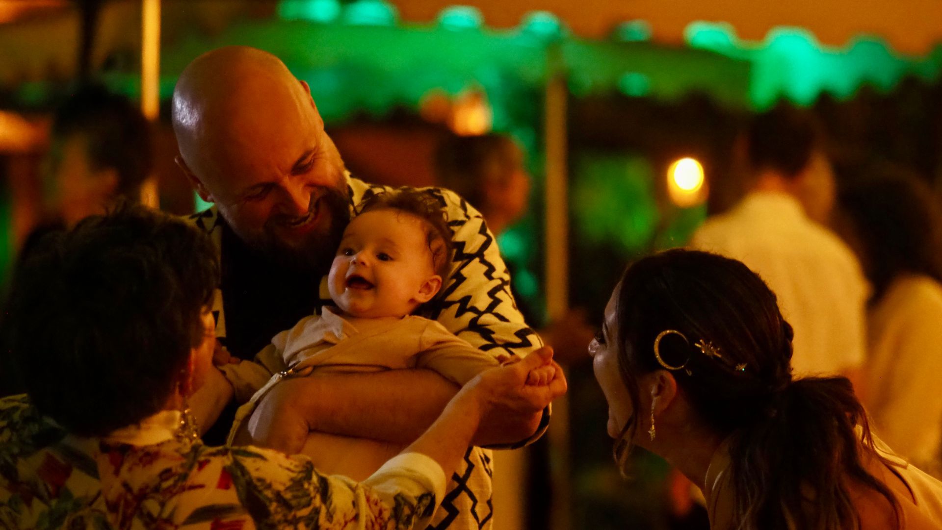 A man holds a smiling baby while two people look on at a warmly lit outdoor gathering.