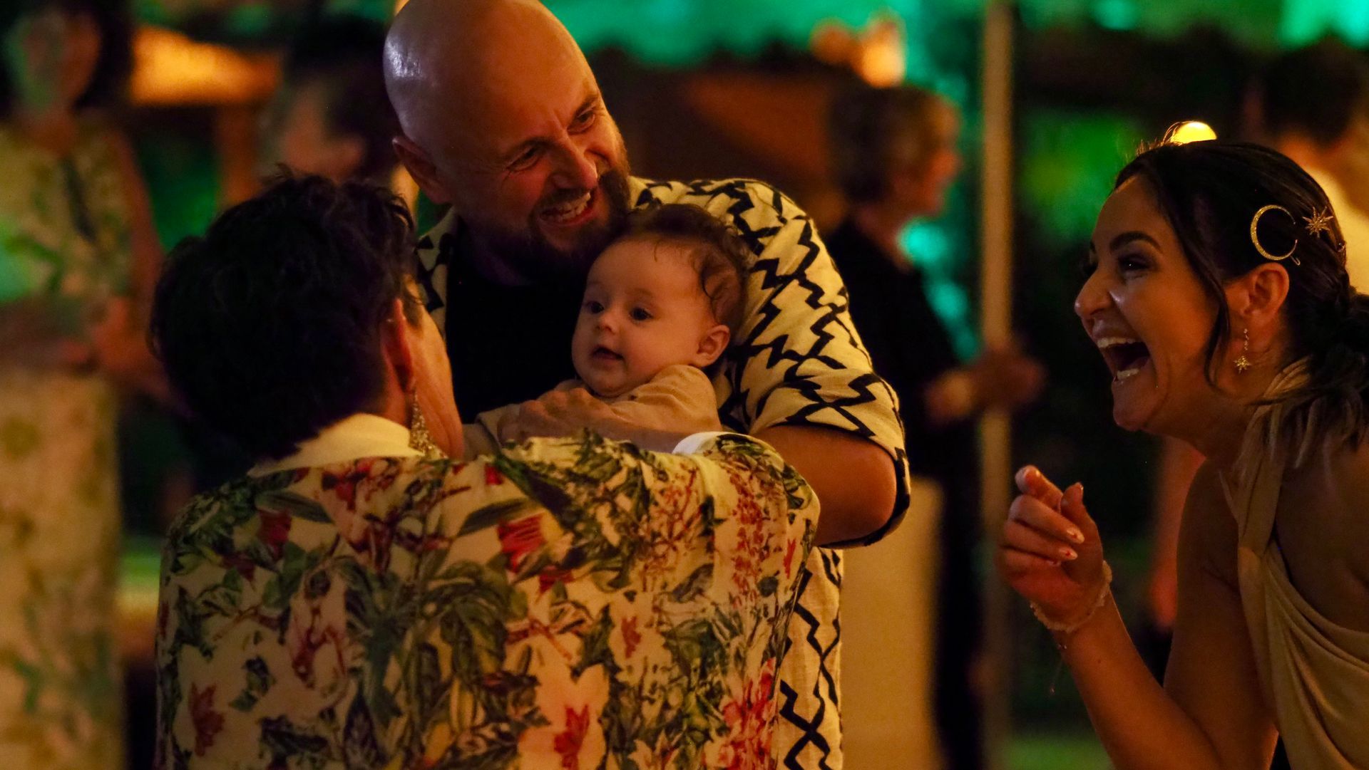 Group sharing a joyful moment at a warmly lit evening gathering, with a baby being held and smiling adults nearby