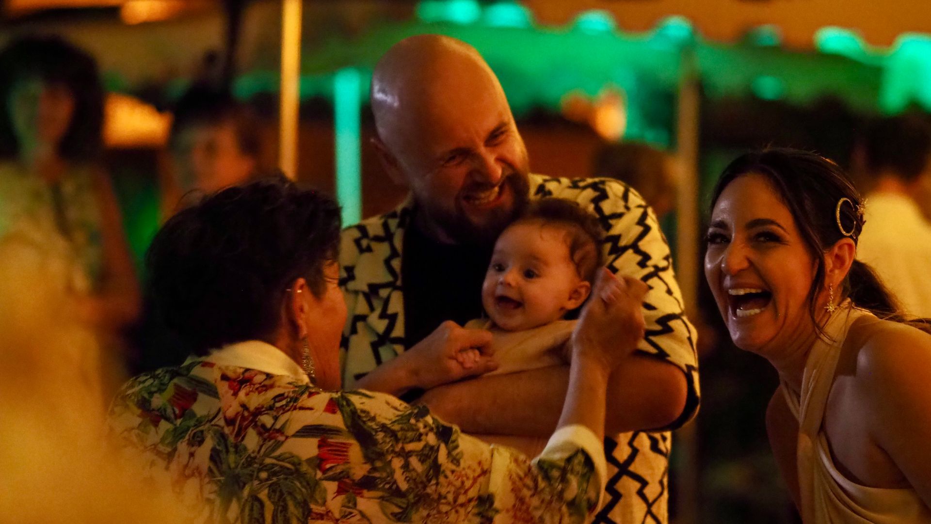 Adults smiling and holding a baby at a warm-lit indoor celebration