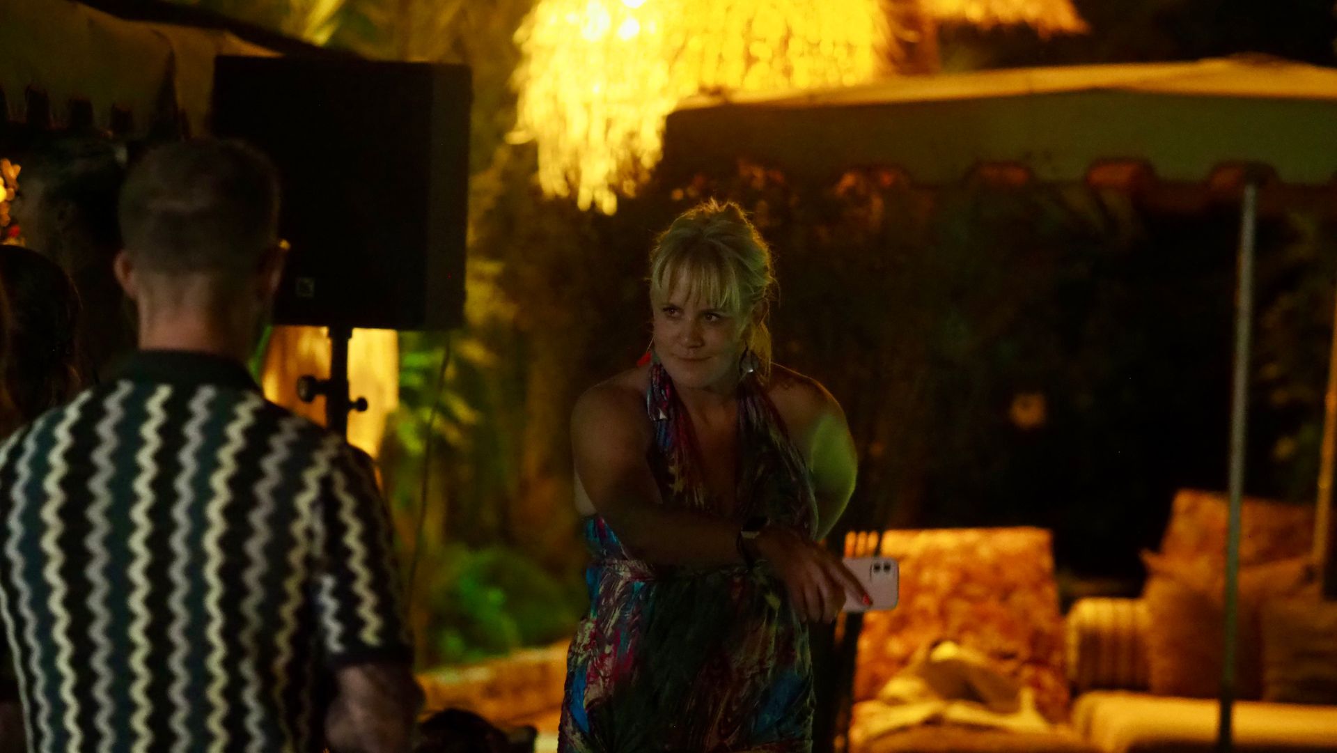 Two people talking in a dimly lit outdoor lounge, with warm lights and a couch in the background