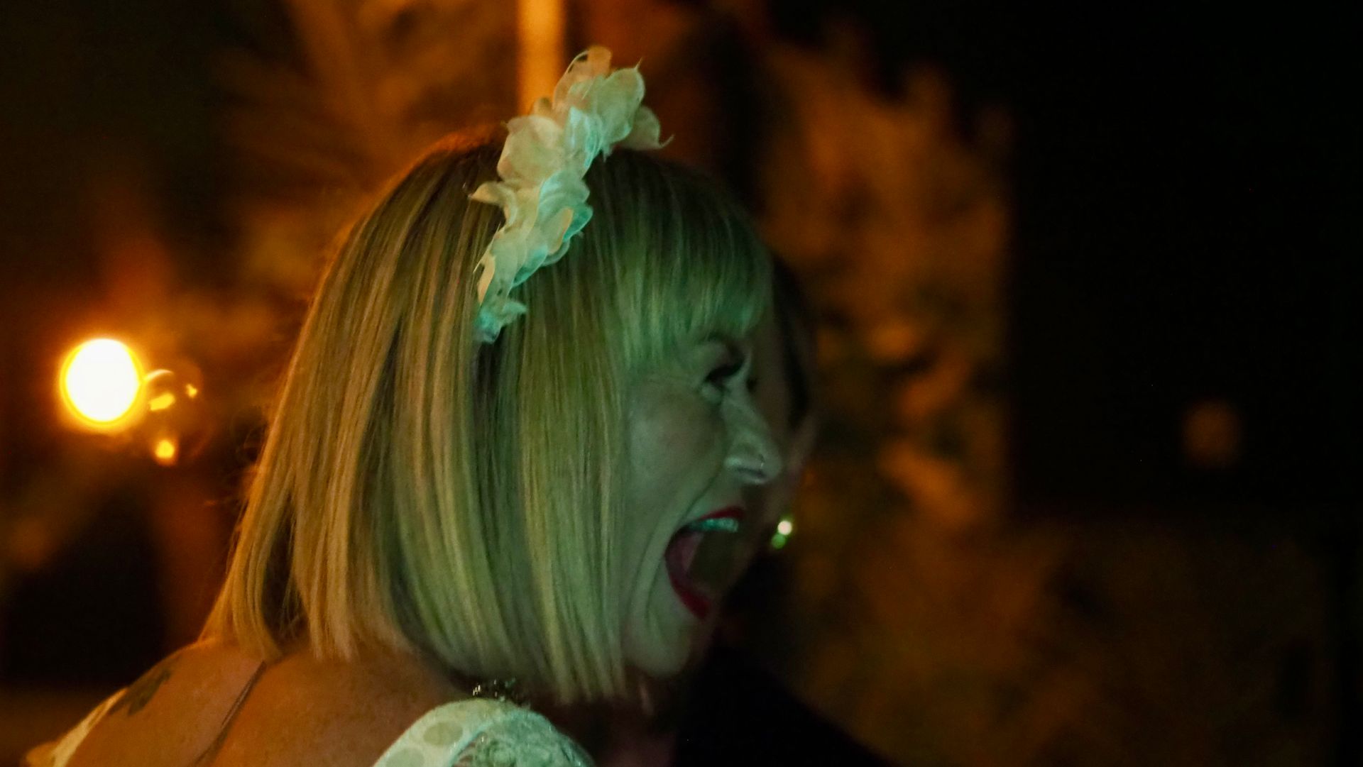 Woman with blonde hair and white headpiece, laughing outdoors at night under warm lights