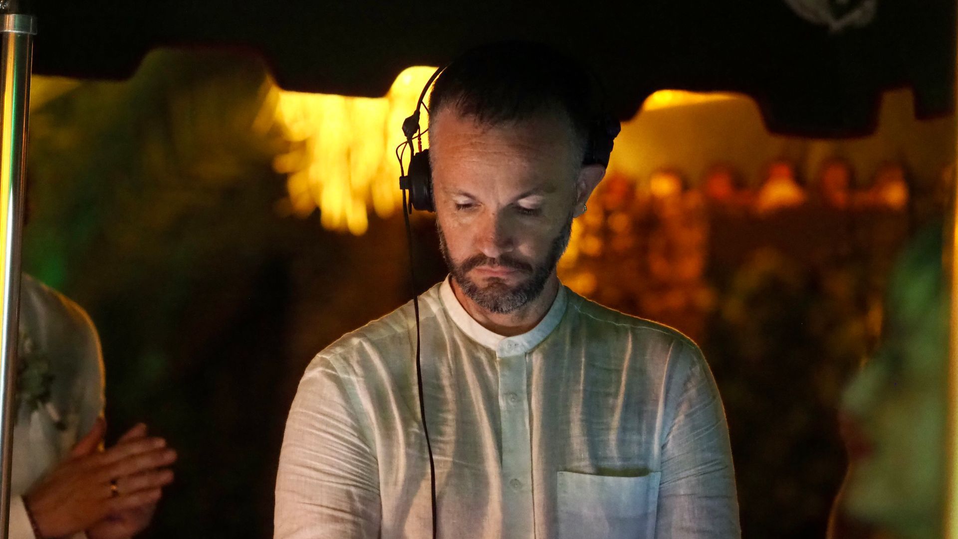 Man in white shirt and headphones looks down under warm yellow lights at a nighttime event