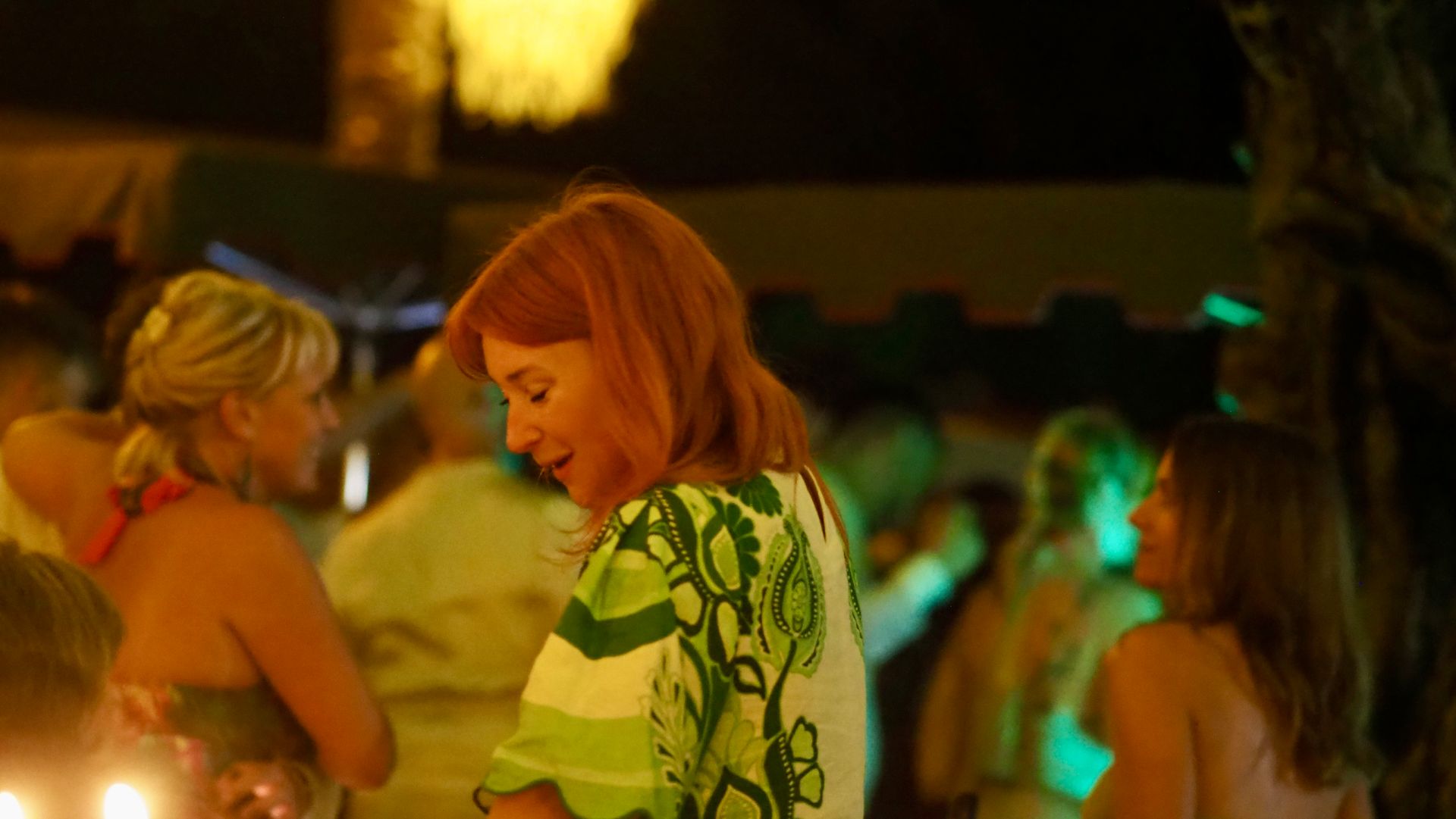 People dancing at a dimly lit party, with a red-haired woman in a green patterned top in the foreground