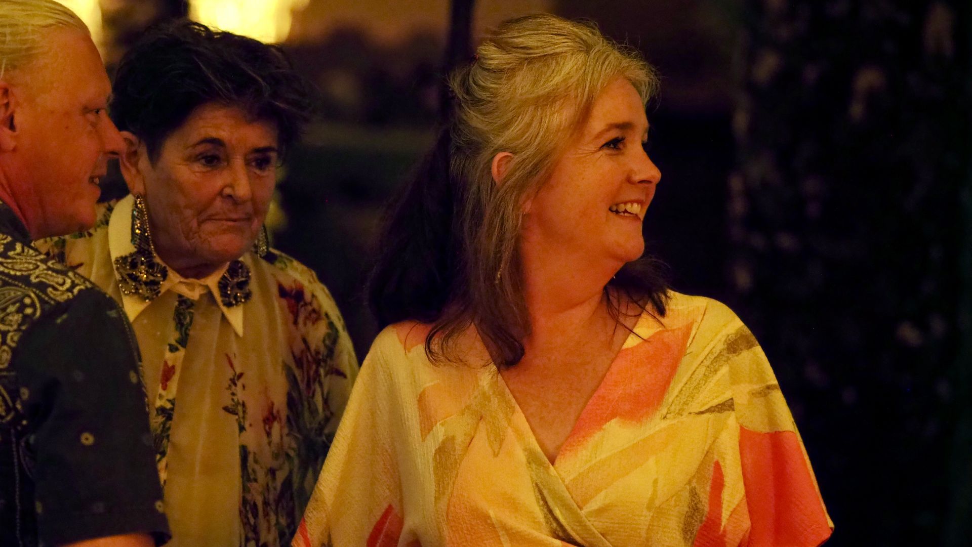 Two people smiling at an evening event, one in a yellow floral blouse and one in a patterned jacket