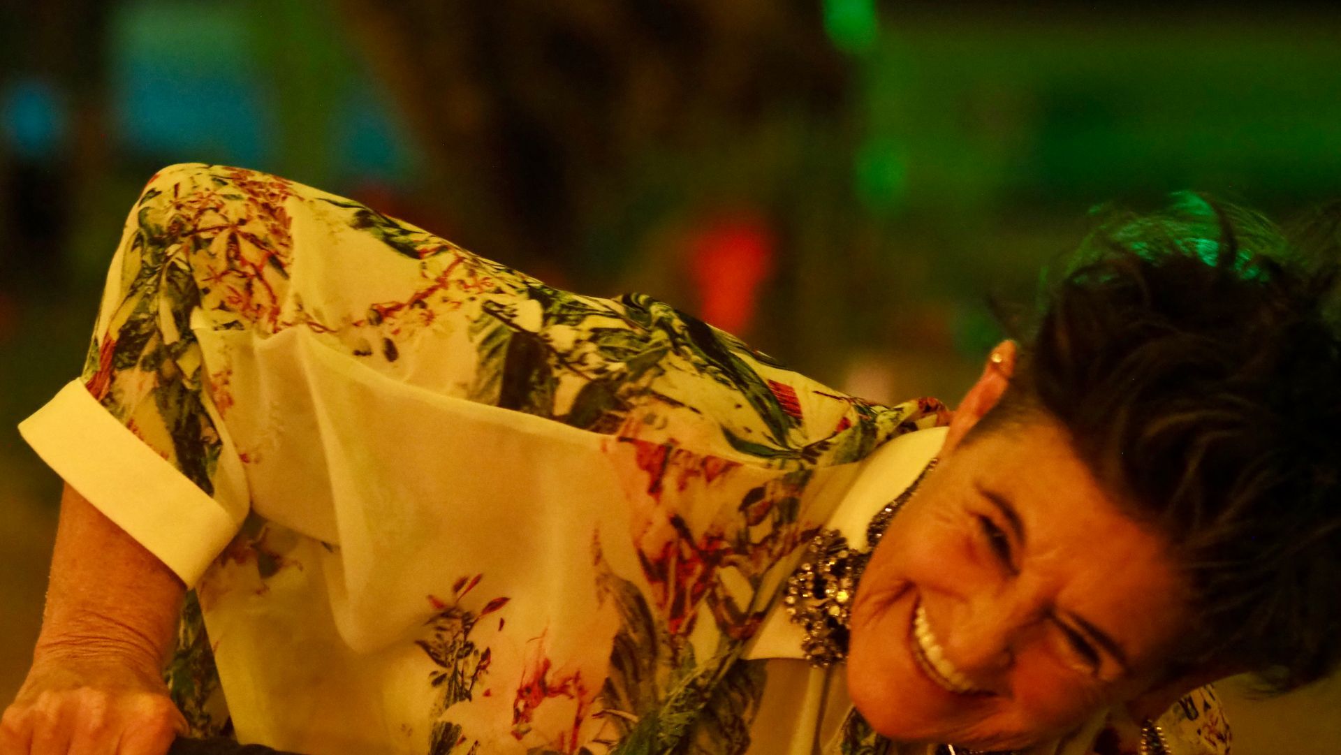 Person in a floral shirt smiling and leaning sideways in warm, colorful lighting