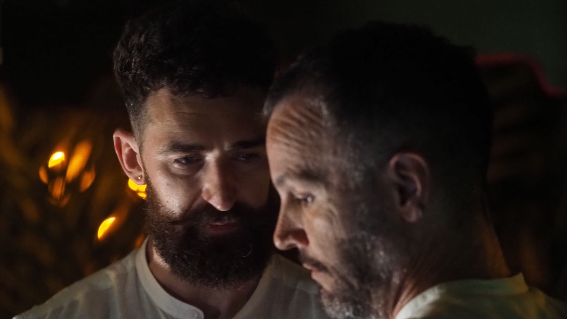 Two men in dim light, one facing the other closely with serious expressions.