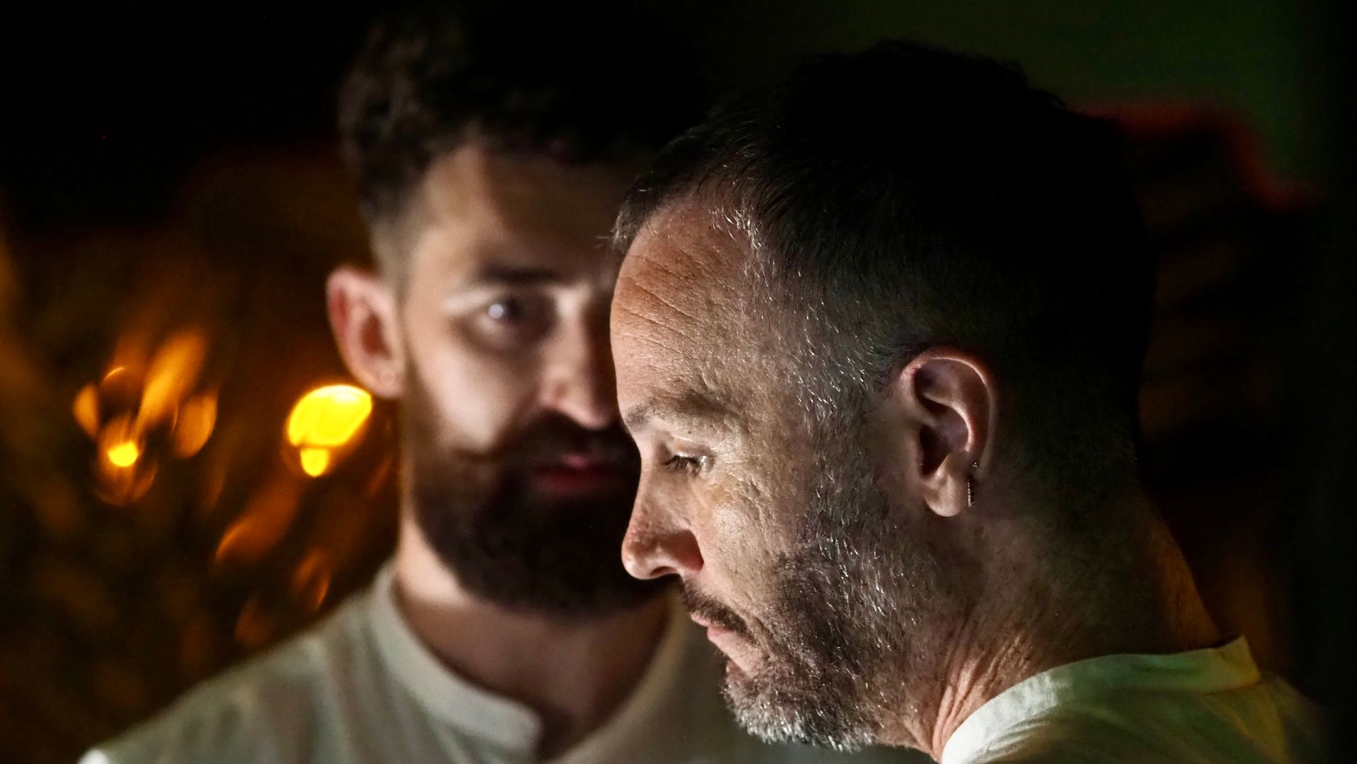 Two men in a dimly lit scene, one in profile with a serious expression and warm lights in the background