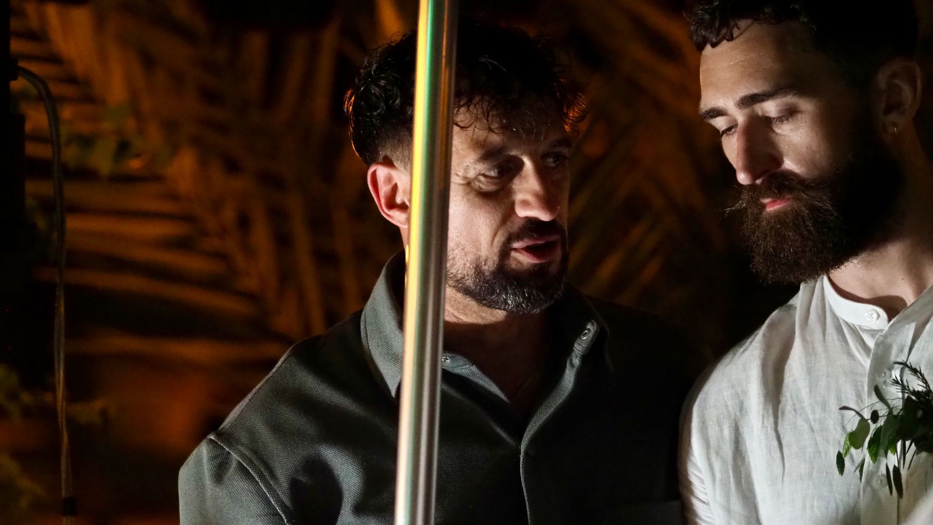Two men standing close together indoors, looking down beside a vertical metal pole in warm light