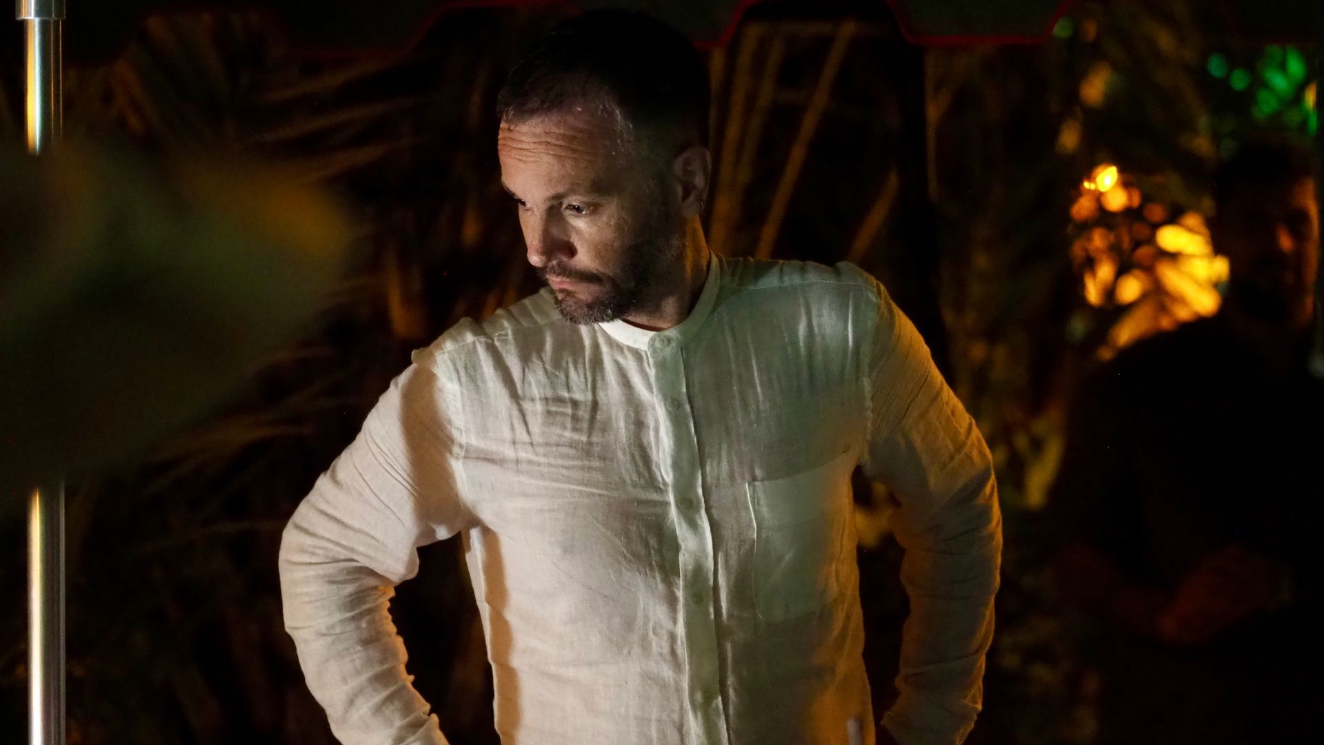 Man in a white shirt standing outdoors at night with warm lights and plants in the background