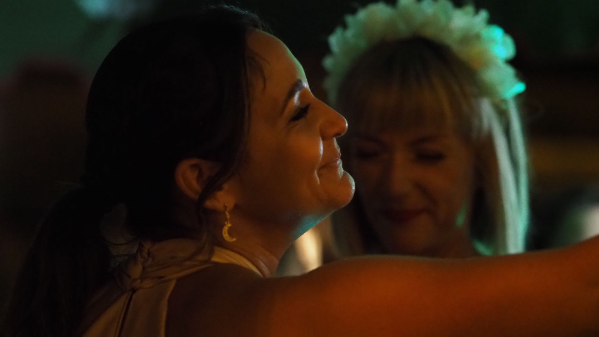Two people dancing closely at a dimly lit party, one smiling in profile with a flower crown behind her.