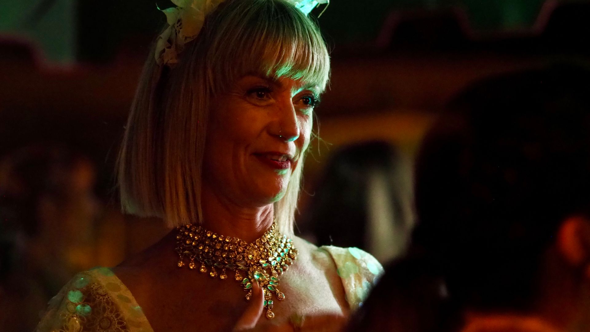 Woman with a blonde bob and jeweled necklace smiling in warm, dim nightclub lighting