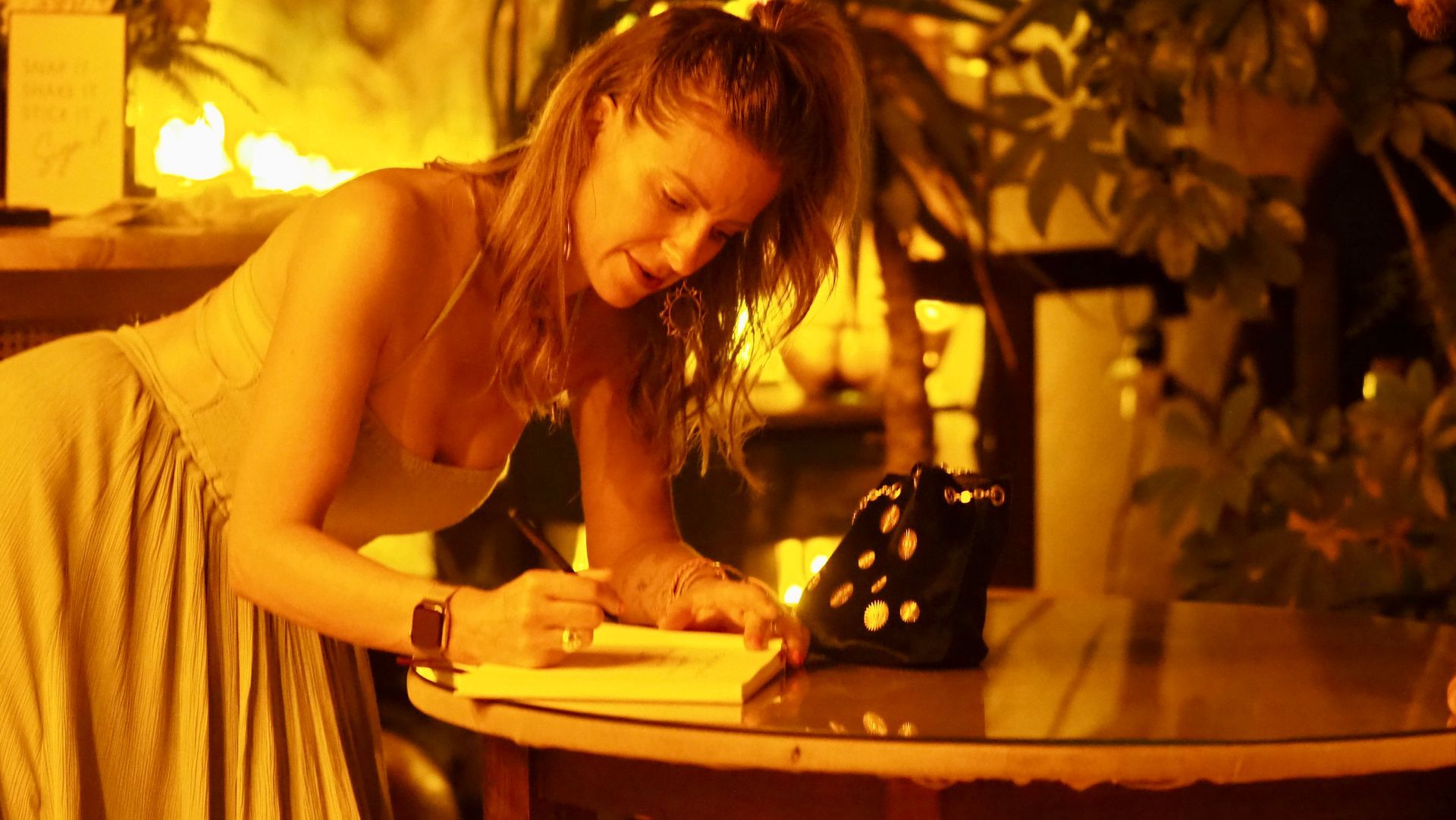Woman writing at a candlelit table, lit by warm orange glow with a telephone nearby.