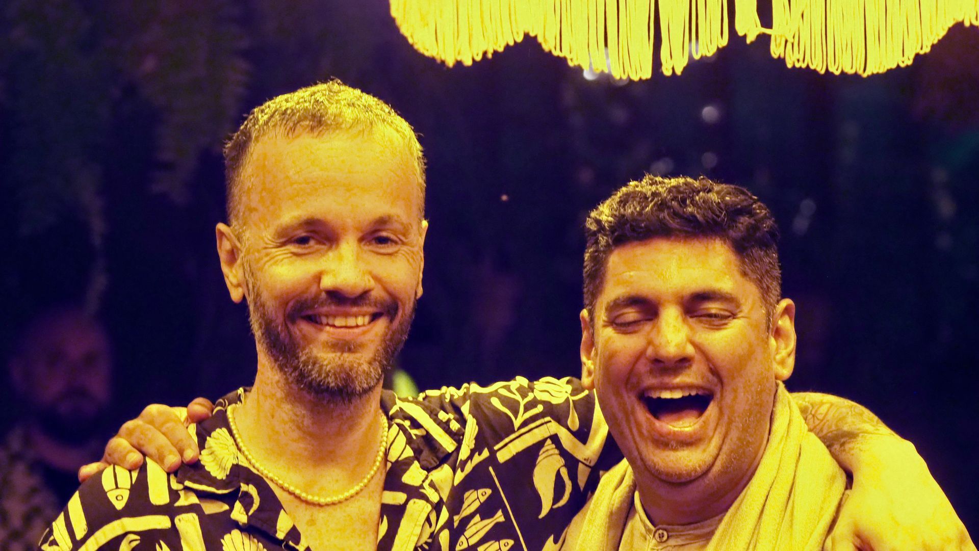 Two smiling people pose shoulder to shoulder under warm yellow lighting, one with an arm around the other.