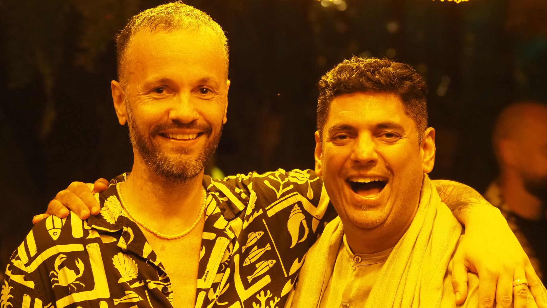 Two smiling people with arms around each other, lit by warm yellow stage lights.
