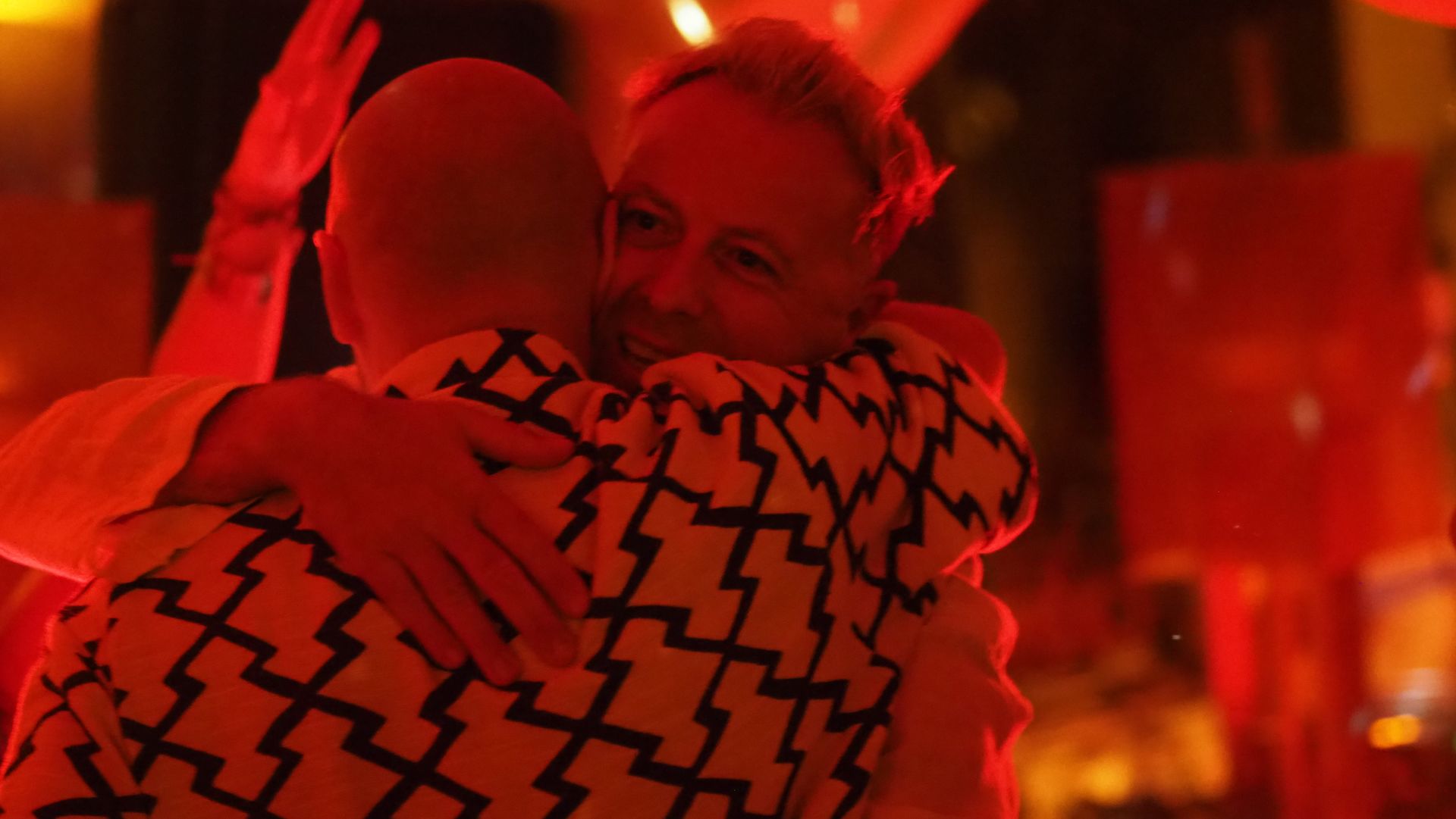 Two people hugging in a red-lit room, one in a black-and-white patterned shirt