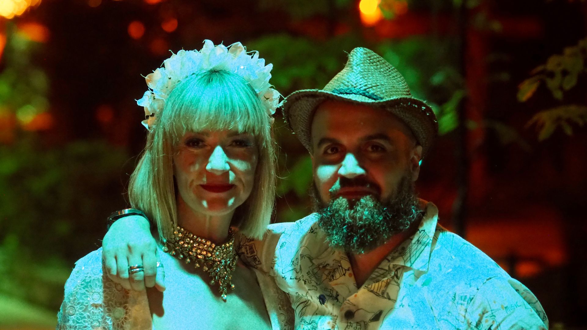 Two people in festive costumes stand together in warm, colorful lighting, one wearing a cowboy hat.