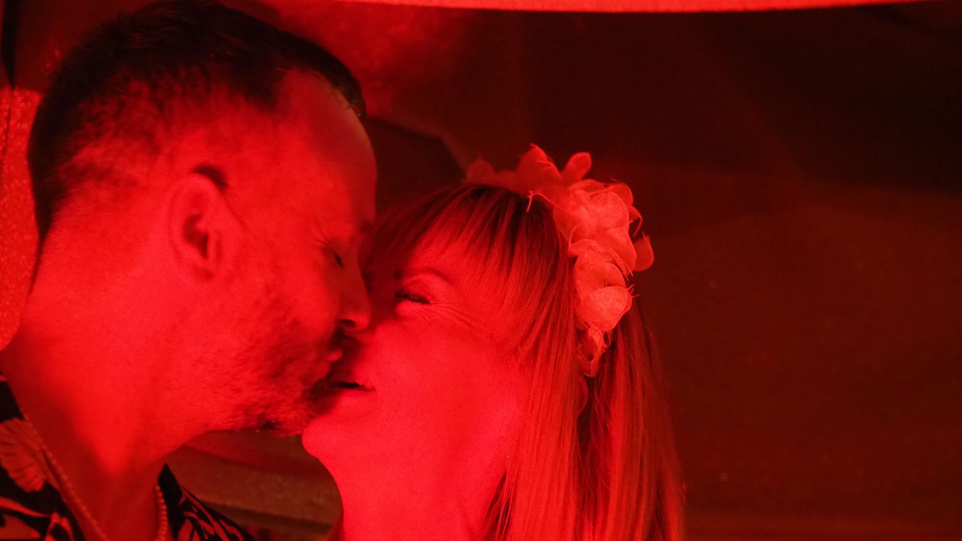 Two people in close profile, leaning in to kiss under red lighting.