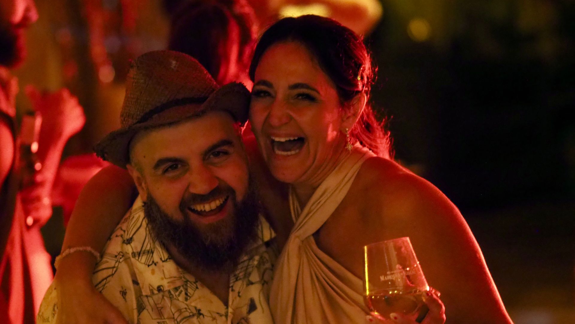 Smiling couple at a warmly lit party, holding drinks and posing close together.