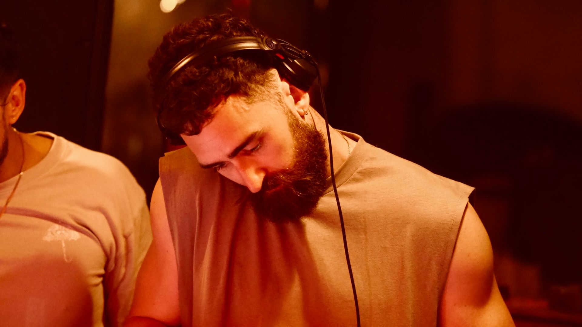 DJ with headphones in warm red lighting, looking down at a mixer or controls