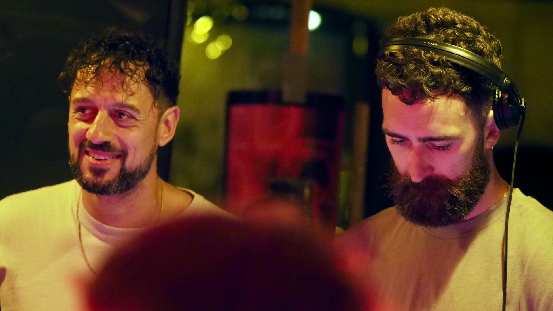 Two DJs at turntables, one smiling and one focused, under warm club lighting