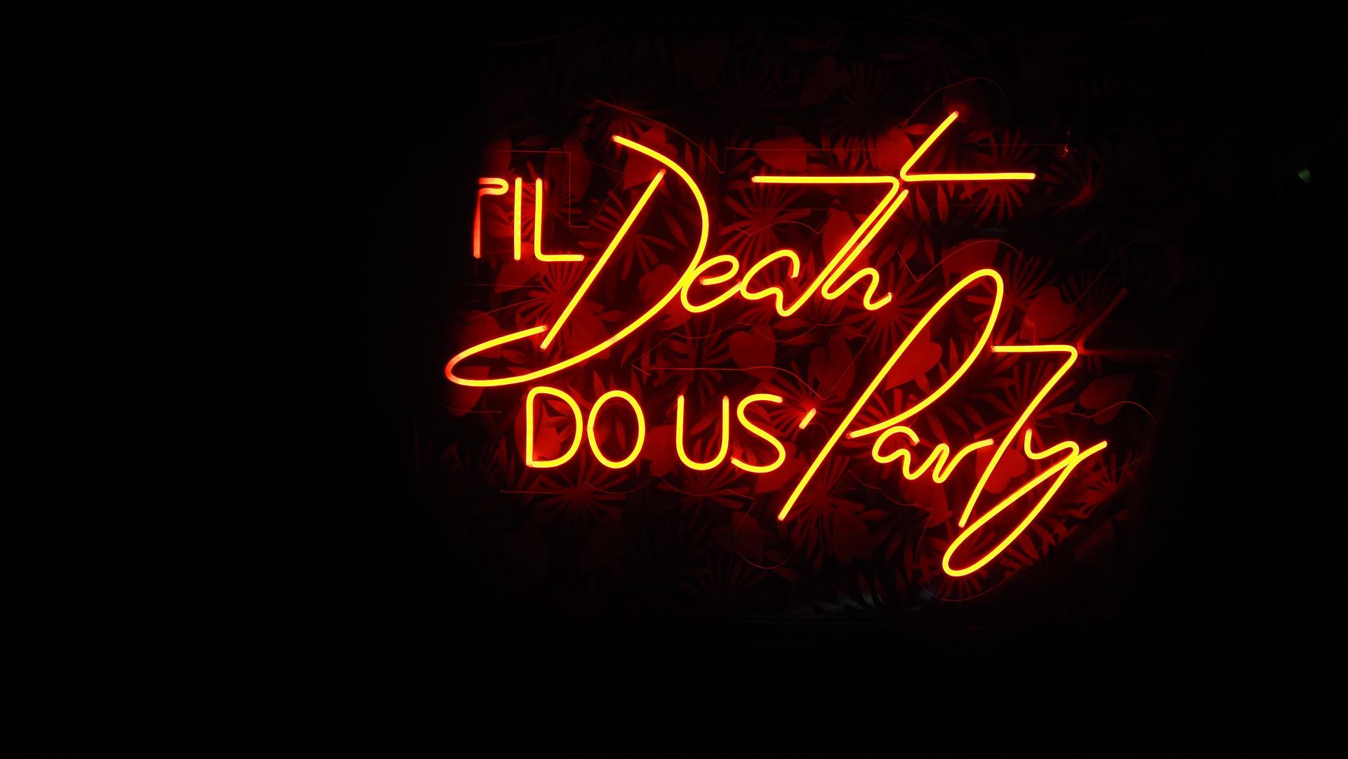 Glowing neon sign reading “Til Death Do Us Party” in red and yellow on a black background