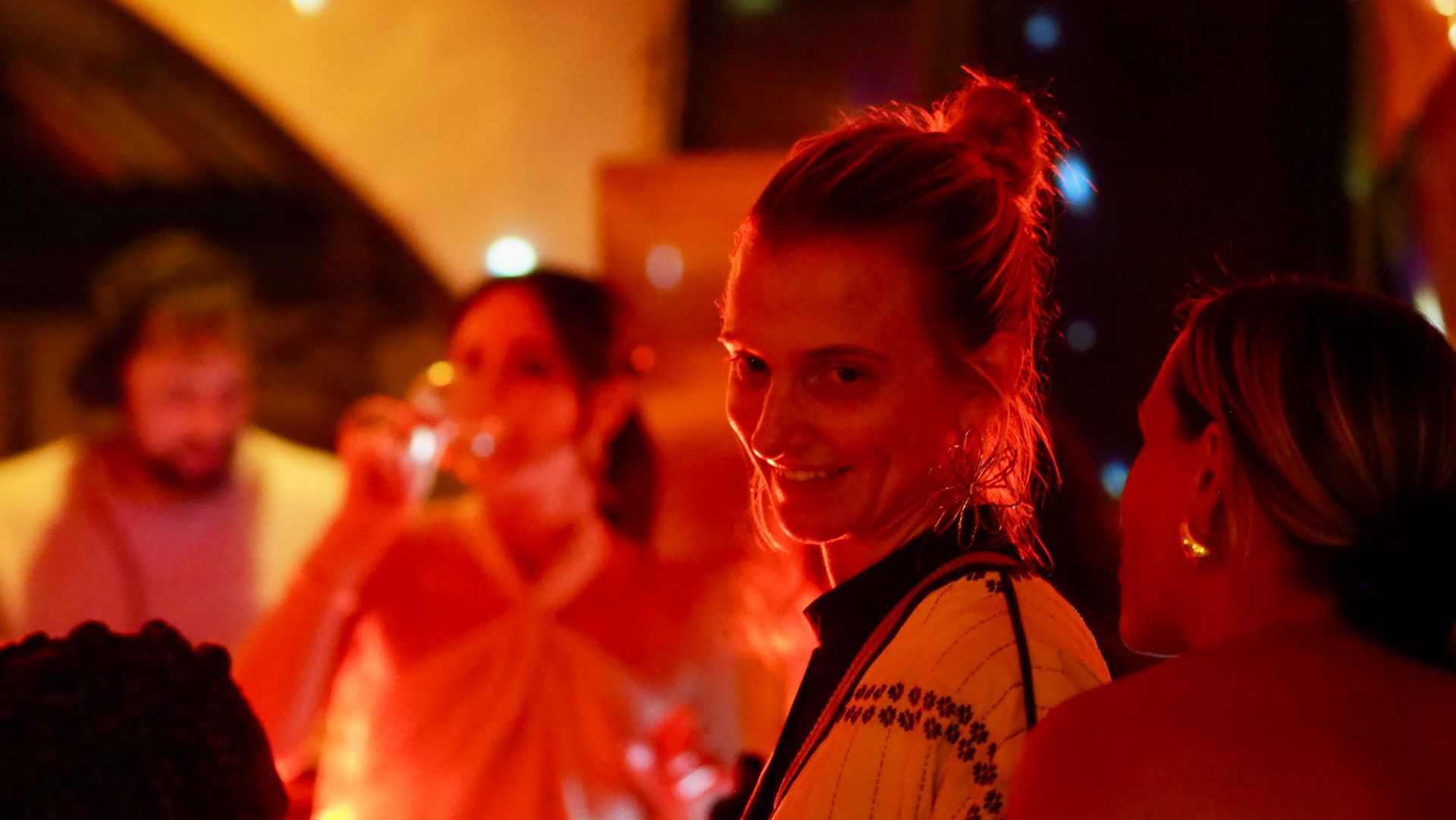 Woman smiling at a warm-lit party, turning toward the camera amid a crowd.