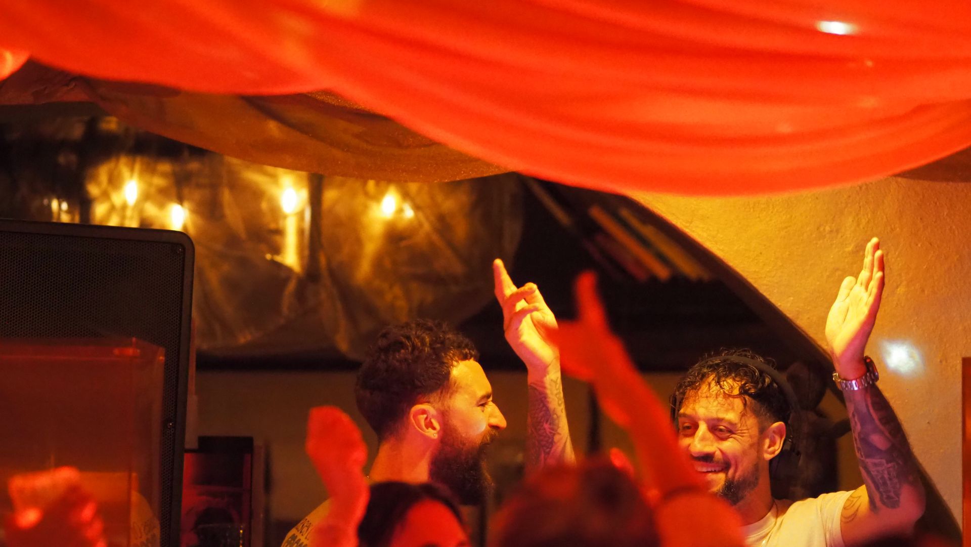 People dancing under orange draped lights at a lively party, with raised hands and warm lighting.