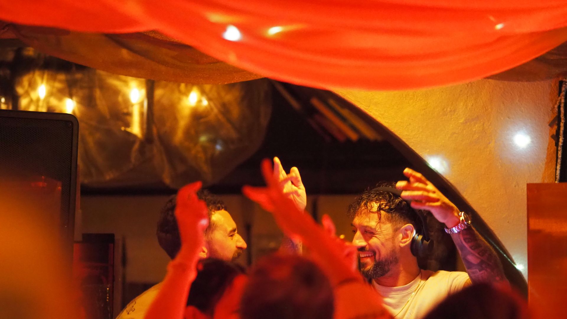 People dancing under red-orange lights at a lively indoor party with raised hands.