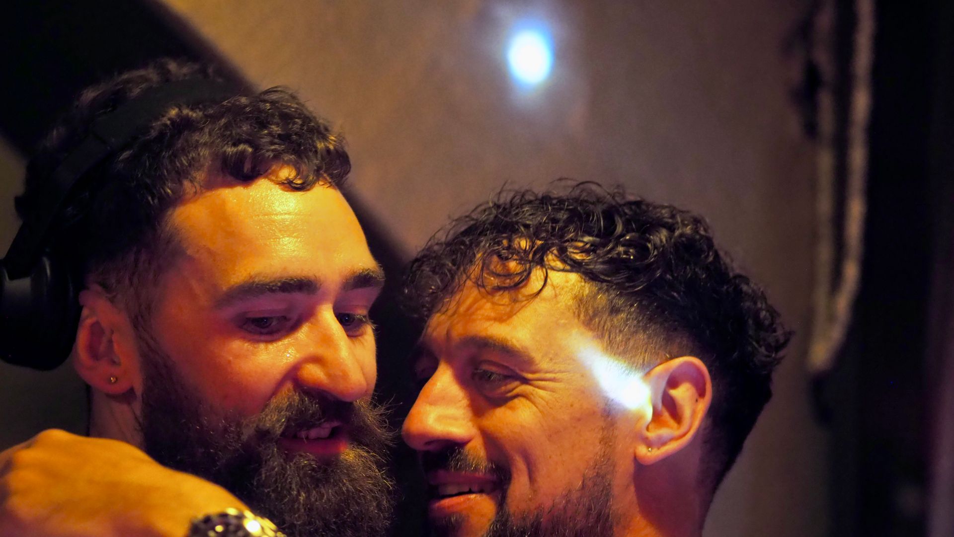 Two smiling men with wet hair lean close together in warm, dim lighting.
