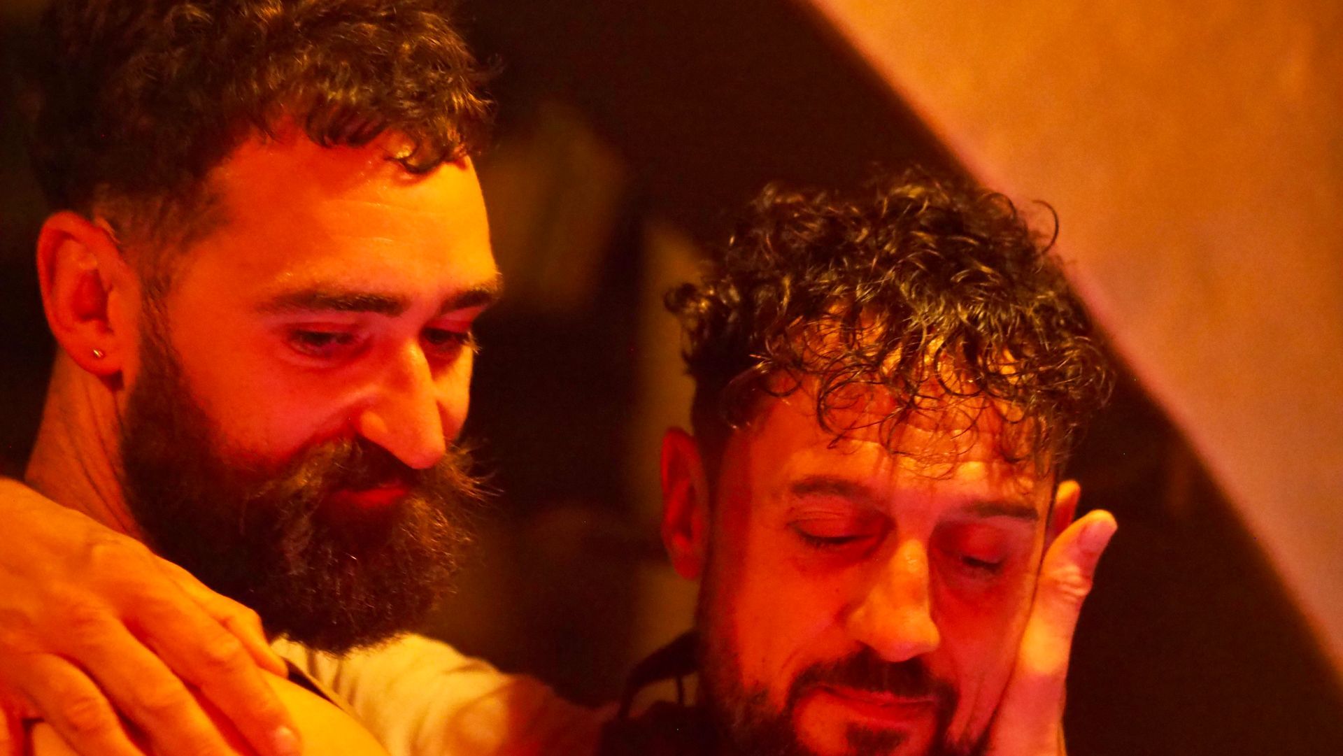 Two men in warm orange light, one smiling and one with eyes closed, standing close together.