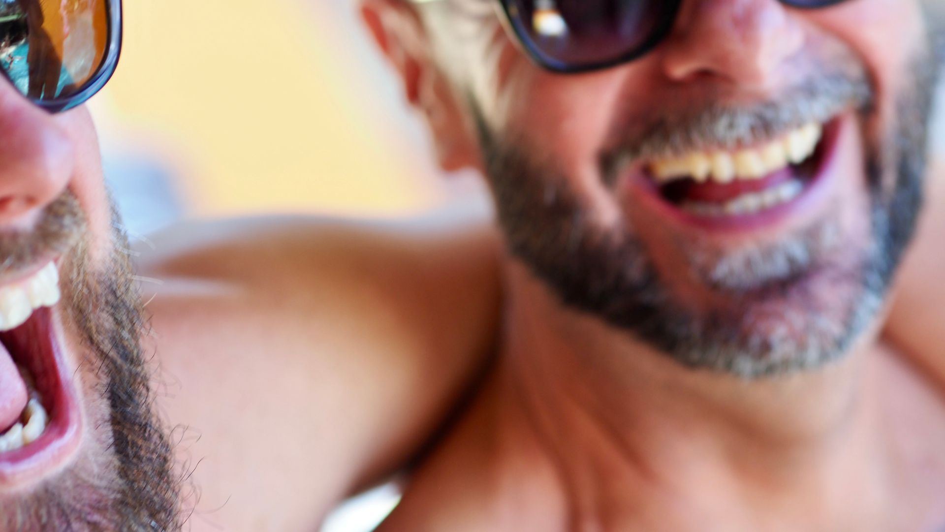 Two smiling people in sunglasses, close together with arms around each other at the beach