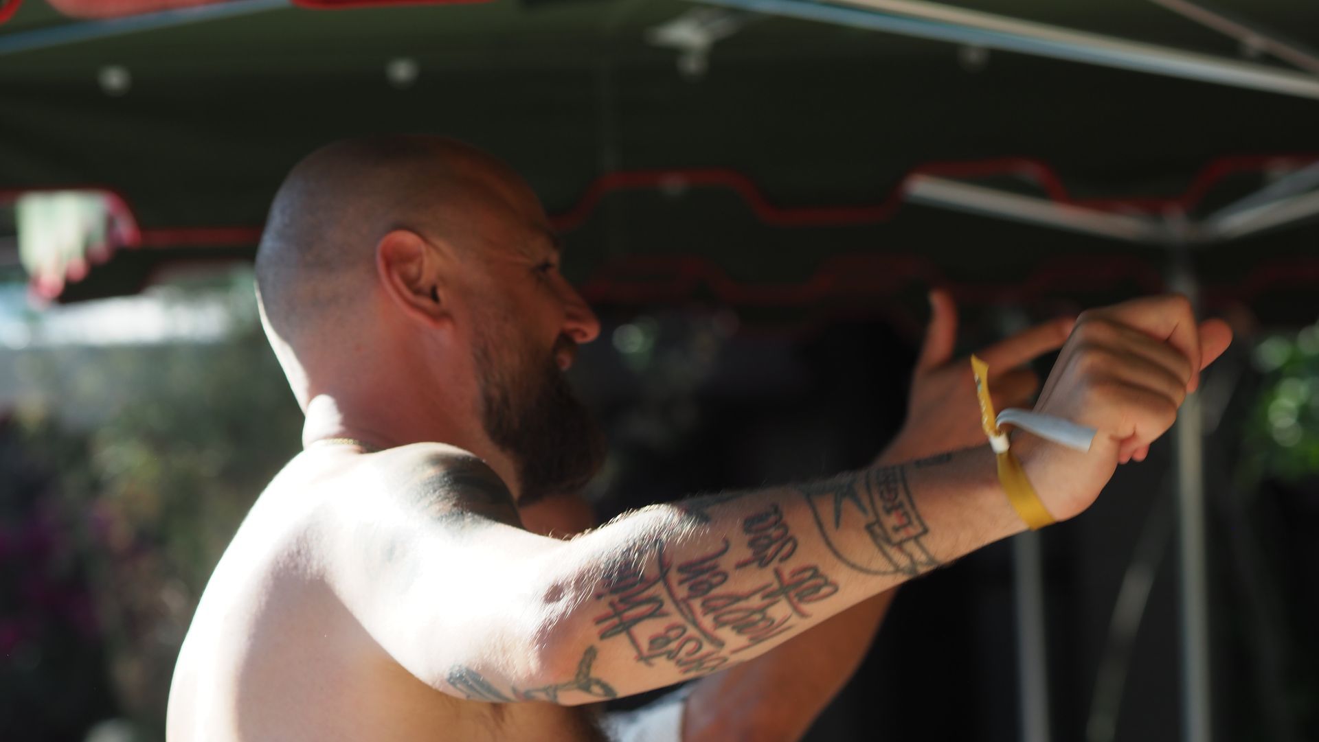 Man with tattooed arm reaching forward under a canopy, facing left in bright sunlight.