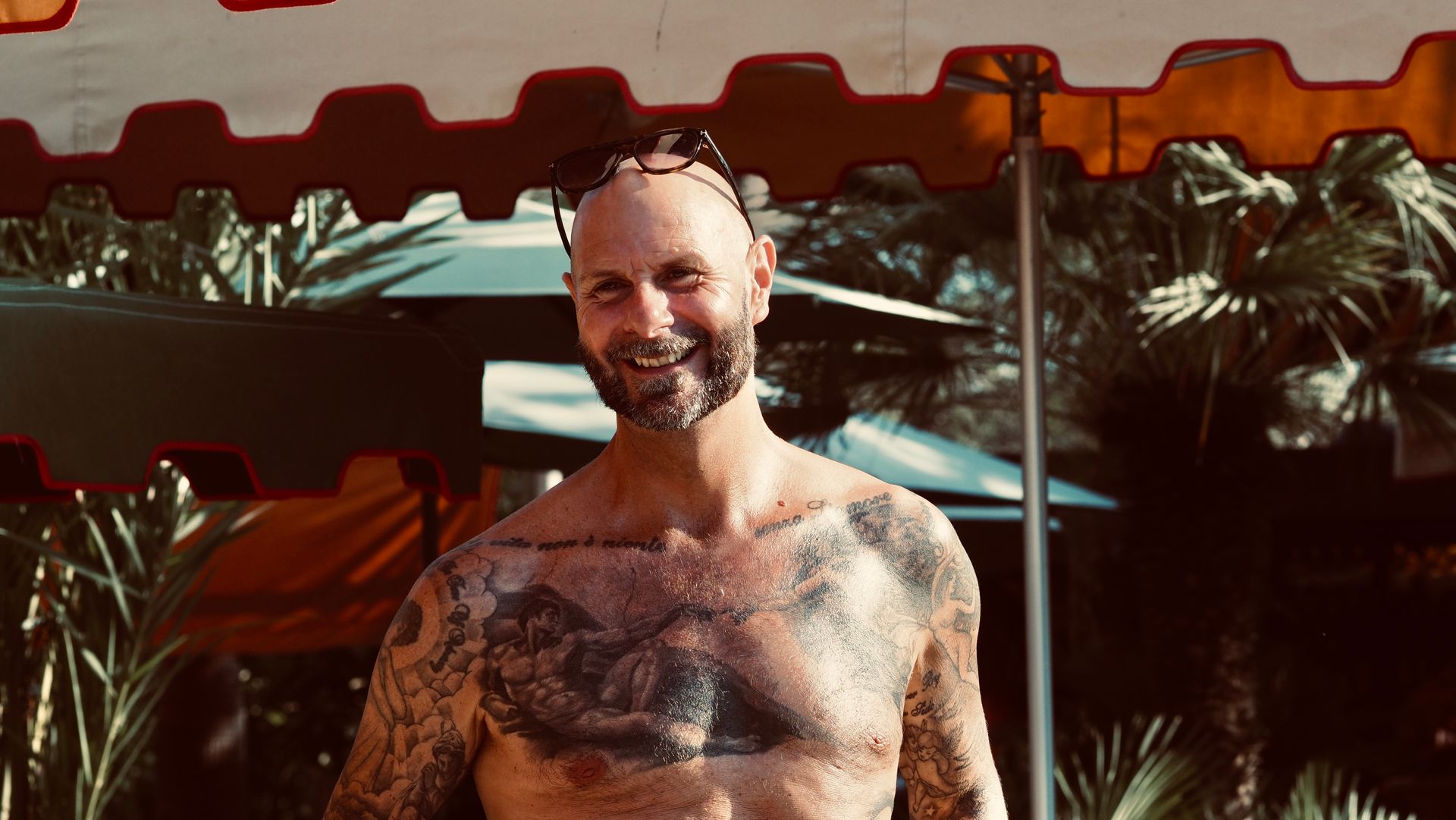 Smiling tattooed man standing outdoors by a red canopy with palm trees in the background